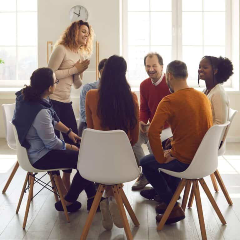   12-Step Group Therapy People sitting in chairs in a circle with woman standing up and speaking to the group